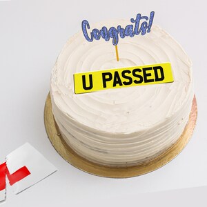 May include: A white cake with yellow frosting and a blue glitter "Congrats!" topper. The cake is decorated with a yellow sign that says "U PASSED".