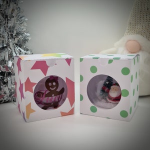 Op de afbeelding: Twee witte cadeaudozen met doorzichtige plastic ramen. De doos aan de linkerkant heeft een roze en wit sterrenpatroon en een peperkoekmannetje erin. De doos aan de rechterkant heeft een groen en wit stippenpatroon en een kerstmanfiguur erin.