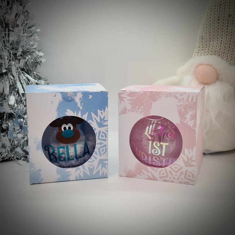 Op de afbeelding: Twee witte dozen met een sneeuwvlokkenpatroon. De doos aan de linkerkant heeft een blauwe achtergrond en een rendier ornament met de naam "Bella" erin. De doos aan de rechterkant heeft een roze achtergrond en een transparant ornament met de tekst "Lily's 1st Christmas" erin.