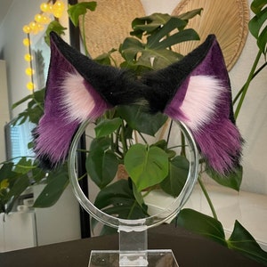 May include: A pair of cosplay cat ears on a clear plastic headband. The ears are black at the tips, transitioning to purple, with a white inner ear. The ears are made of a soft, furry material.