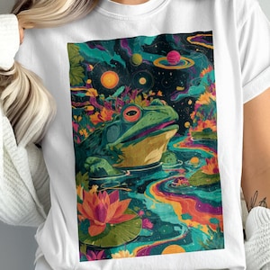 May include: White t-shirt featuring a vibrant, psychedelic illustration of a frog in a pond with planets and colorful water. The artwork includes a frog, lily pads, and a cosmic background, creating a unique and artistic design.