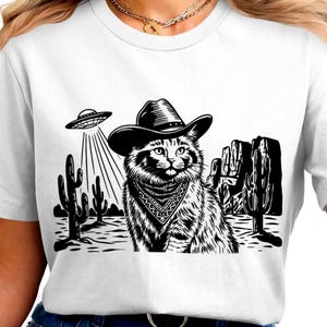 Puede incluir: Camiseta blanca con un gráfico en blanco y negro. El gráfico representa un gato con sombrero de vaquero y pañuelo, con un OVNI que lo ilumina. Escena desértica con cactus y formaciones rocosas.