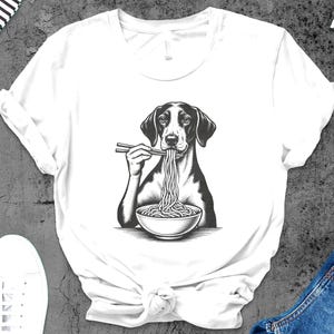 May include: White t-shirt featuring a black and white illustration of a dog eating noodles with chopsticks from a bowl. The dog has black ears and a black and white face. The shirt is on a gray surface with white shoes and blue jeans.