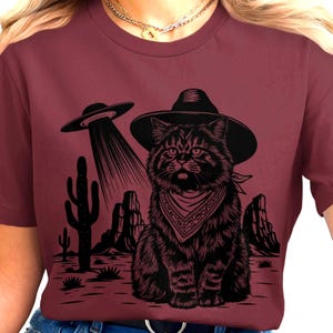 Puede incluir: Camiseta burdeos con un diseño gráfico negro de un gato con sombrero de vaquero y pañuelo, con un OVNI. El gato es el foco central del diseño, en un paisaje desértico.