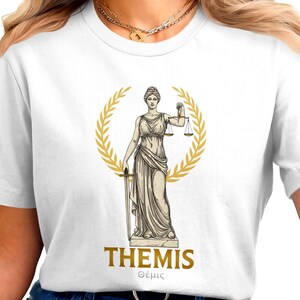 Puede incluir: Camiseta blanca con un diseño dorado de Temis, la diosa griega de la justicia, sosteniendo una balanza y una espada, rodeada por una corona de laurel dorada. La palabra "THEMIS" está impresa en dorado, con la palabra griega debajo.