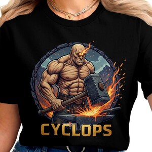 May include: Black t-shirt featuring a muscular figure with a glowing eye, holding a hammer over an anvil. The word "CYCLOPS" is displayed in gold letters below the illustration. The design is set against a backdrop of flames and a mountain landscape.