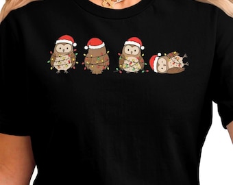 May include: Black t-shirt featuring a festive owl design. Four cartoon owls are depicted, each wearing a Santa hat and wrapped in Christmas lights. One owl is lying on its back. A fun, holiday-themed graphic.