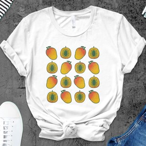 以下が含まれることがあります： マンゴーのイラストが繰り返されるパターンの白いTシャツ。マンゴーは黄色、オレンジ、緑色で、ヴィンテージスタイルです。白いスニーカーとデニムジーンズでスタイリングされています。