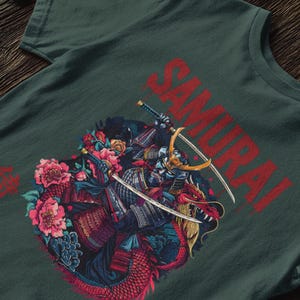 May include: Dark green t-shirt featuring a detailed illustration of a samurai warrior with two swords, riding a red dragon. The word "SAMURAI" is printed in red across the top. Pink and blue flowers surround the warrior and dragon.