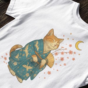 May include: White t-shirt featuring a whimsical illustration of a ginger cat wearing a blue kimono with crane designs. The cat holds a fan and is surrounded by cherry blossoms and a crescent moon.