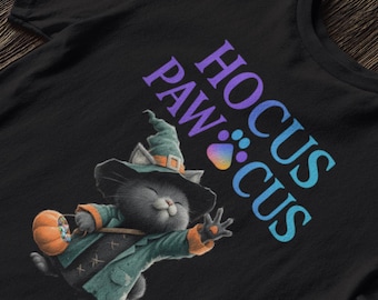 Op de afbeelding: Zwart t-shirt met een cartoonkat verkleed als heks, met een pompoen. De kat draagt een groene hoed en jas. De tekst "Hocus Paws Cus" is gedrukt in paars en blauw.