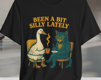 Op de afbeelding: Zwart T-shirt met een witte gans en een zwarte kat met een bril. De tekst "Been a bit silly lately" staat op het shirt gedrukt.