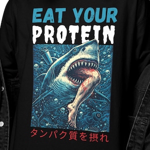 Może przedstawiać: Czarna koszulka z długim rękawem z grafiką rekina jedzącego nogę, z napisem "EAT YOUR PROTEIN" w kolorze niebieskim i białym. Japoński tekst poniżej. Koszulka jest noszona przez osobę.