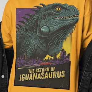 Op de afbeelding: Een mosterdgeel t-shirt met een grafische illustratie van een grote groene leguaan die boven een stadssilhouet uittorent. De tekst "THE RETURN OF IGUANASAURUS" staat in het geel onderaan het rechthoekige ontwerp, dat een rode auto bevat.