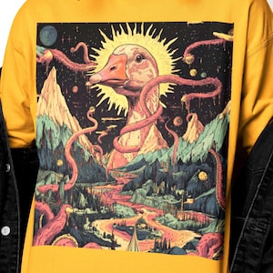 Puede incluir: Camiseta de manga larga de color amarillo mostaza con una ilustración surrealista de un ganso con un halo solar, rodeado de montañas, un río y elementos cósmicos. La obra de arte está realizada en colores vibrantes, creando un diseño único y llamativo.