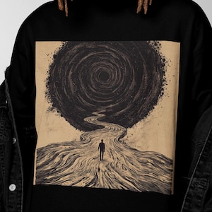 Może przedstawiać: Czarny t-shirt z okrągłym dekoltem z surrealistyczną ilustracją. Grafika przedstawia postać idącą ścieżką w kierunku wirującego wiru. Projekt jest w odcieniach brązu i czerni, tworząc uderzający kontrast. Koszulka jest wykonana z miękkiego materiału.
