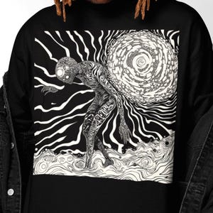 Puede incluir: Camiseta negra de manga larga con un gráfico en blanco y negro de una figura que lleva un orbe arremolinado. La obra de arte tiene un estilo ilustrativo detallado con líneas onduladas y un tema surrealista.