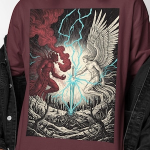 Könnte beinhalten: Burgunderfarbenes Langarmshirt mit einer Grafik, die einen Kampf zwischen einem Engel und einem Dämon darstellt. Der Engel hat große weiße Flügel und der Dämon ist rot. Ein blaues Schwert befindet sich zwischen ihnen, mit Blitzen im Hintergrund.