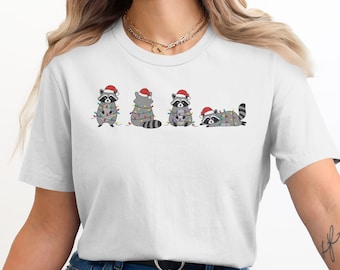 Può includere: T-shirt bianca con una grafica di quattro procioni con cappelli di Babbo Natale e avvolti in luci natalizie. I procioni sono in diverse pose. Il disegno è centrato sulla maglietta.