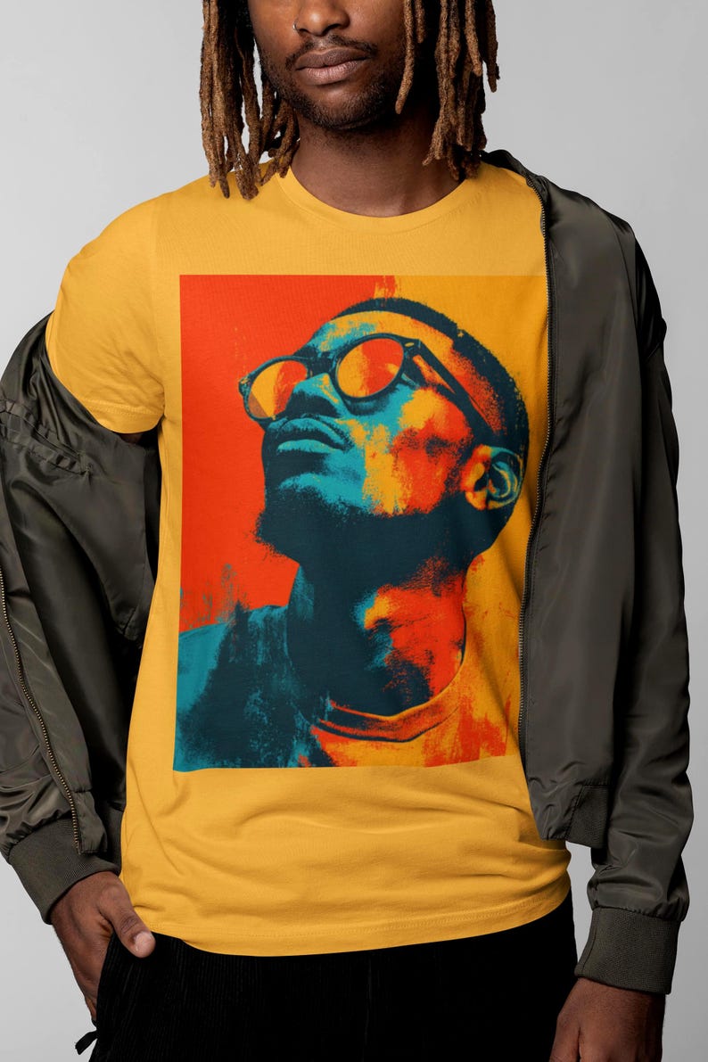 May include: Mustard yellow t-shirt with a pop art portrait of a person wearing glasses, in orange and blue tones. The person is looking upwards. The shirt is worn with a dark green jacket.