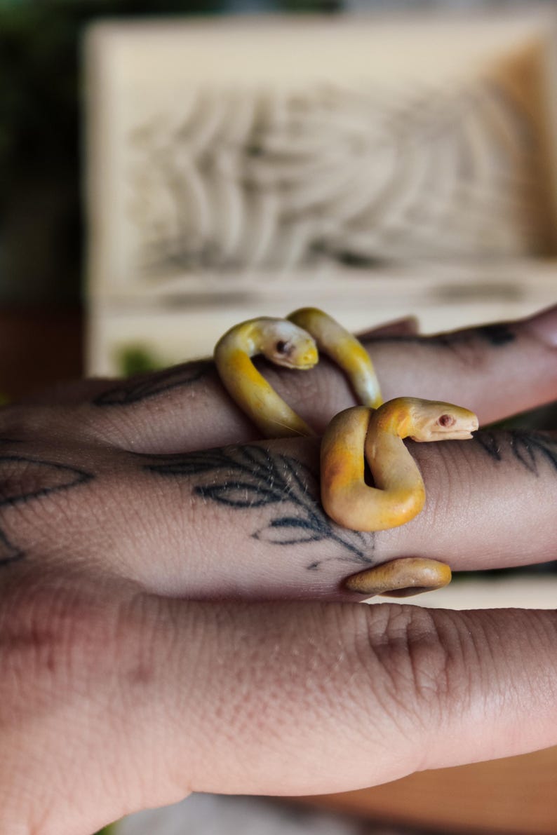 May include: A ring shaped like a coiled snake with a smooth, matte finish in shades of pale yellow and orange. The snake features a detailed head with visible eyes and a curved body that wraps around the finger, creating a continuous, fluid form.