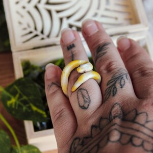 May include: A ring shaped like a coiled snake with a smooth, pale yellow and white gradient finish. The snake's body wraps around the finger, with the head resting against the tail, creating a continuous, circular band design for the finger.