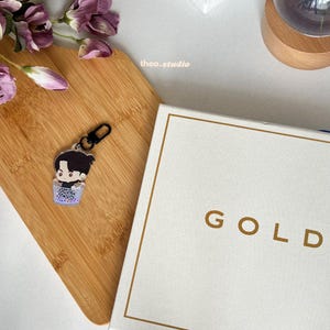 Puede incluir: Un llavero transparente con una ilustración de dibujos animados de una persona con cabello negro que lleva una camisa negra y un cuello blanco. La persona sostiene un pequeño objeto blanco en sus manos. El llavero está sobre una superficie de madera.