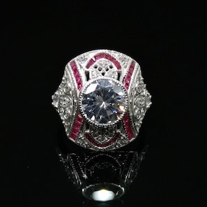 Puede incluir: Un anillo de plata con una gran piedra redonda central transparente rodeada de piedras más pequeñas. El anillo está acentuado con una banda de piedras rojas.