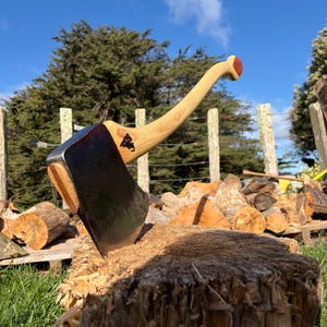 Op de afbeelding: Een stalen bijlkop met een donkere, verweerde afwerking die in een grote houten blok steekt. De bijl heeft een gebogen, lichtgekleurde houten steel met een roodbruin accent bij de knop en een klein zwart logo op de hals.