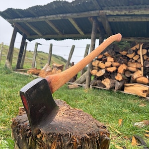 Puede incluir: Un hacha de tala con cabeza de metal oscuro que presenta un detalle rojo en el talón y un mango de madera curvado. El hacha está incrustada en un tocón de madera circular y desgastado, mostrando una veta texturizada y un filo metálico.