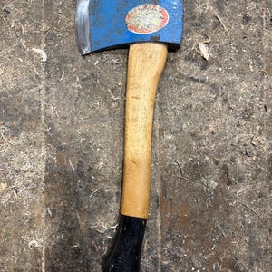 Op de afbeelding: Een blauwe bijl met een houten steel en een zwarte grip. De bijlkop heeft een zilveren blad en een ronde sticker. De bijl ligt op een houten oppervlak bedekt met houtkrullen.