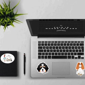 May include: A laptop computer with three stickers on it. The stickers are of a woman with dogs, a black and white dog with the name "Winston" and a brown and white dog with the name "Dash".