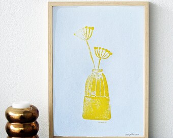 May include: A framed art print featuring a yellow floral design in a vase against a light blue background. The print is in a natural wood frame. The artwork is signed in the bottom right corner.
