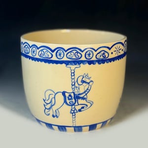 May include: A cream-colored ceramic planter with a blue carousel horse design. The planter has a blue decorative border at the top and a blue striped base. The carousel horse is painted in blue.