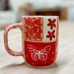 May include: Hand-painted ceramic mug with a patchwork design. The mug features a red base with a white butterfly, hearts, and flowers. The handle is pink with white polka dots. A unique and colorful addition to any kitchen.