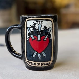 May include: Black ceramic mug featuring a tarot card design. The card depicts a red heart pierced by three swords, with the Roman numeral III at the top and the words "THREE OF SWORDS" at the bottom. The mug has a curved handle.