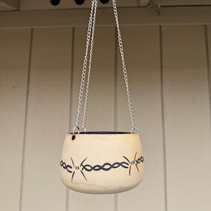 May include: A beige ceramic hanging planter with a black barbed wire design. The planter is suspended by a silver chain. The planter is designed to hold plants and flowers.