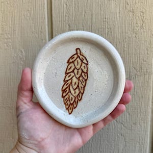 May include: A small, round, off-white ceramic spoon rest with a brown hop design. The spoon rest has a speckled texture and a slightly raised edge. The hop design is hand-painted in shades of brown.