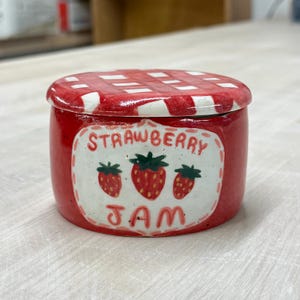 May include: A small, round, red ceramic jar with a lid featuring a red and white checkered pattern. The jar has a label that reads "STRAWBERRY JAM" with illustrations of strawberries.
