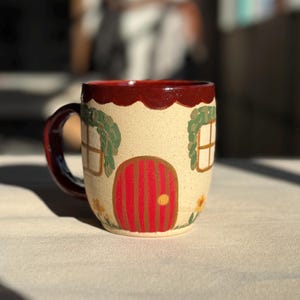 May include: A whimsical ceramic mug featuring a house design. The mug has a cream-colored base with a red door, windows, and a scalloped rim. Green foliage and yellow flowers add detail. The handle and interior are a deep red.