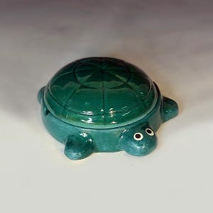 Puede incluir: Caja de baratijas de cerámica con forma de tortuga en verde azulado brillante. La tortuga tiene un diseño de caparazón detallado y pequeños ojos con borde blanco. La tapa encaja perfectamente, ideal para guardar objetos pequeños.