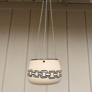 May include: Cream-colored hanging planter with a black chain link design around the middle. The planter is suspended by three silver chains. The rim of the planter is black.