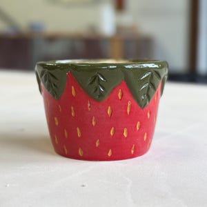 May include: A ceramic strawberry-themed planter. The planter is red with yellow seed details and a green top. The planter is designed to resemble a strawberry, perfect for small plants or succulents.