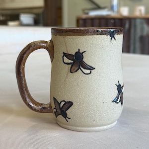 May include: A speckled beige ceramic mug featuring a hand-painted pattern of dark brown insects with black outlines. The mug has a thick, curved handle glazed in a glossy dark brown, matching the rim, and a textured, matte-finish body.
