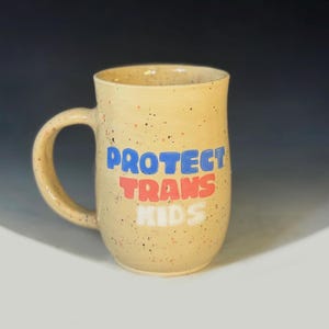 May include: A speckled, beige ceramic mug with a curved handle. The mug features the text "PROTECT TRANS KIDS" in blue, red, and white block letters. The mug is set against a neutral background.