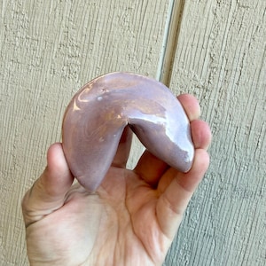 May include: A ceramic fortune cookie in a lavender color with a glossy finish. The cookie is held in a hand, showcasing its curved shape and smooth texture. The background is a light-colored wooden surface.