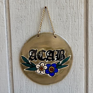 May include: Round, gold-colored ceramic wall hanging with the letters "ACAB" in black gothic font. The sign features a white and blue flower design with green leaves. A gold chain is attached for hanging.