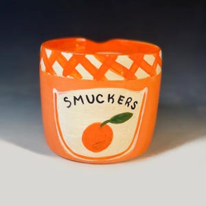 May include: Orange ceramic jar with a white label featuring the word "SMUCKERS" and an orange illustration. The jar has a decorative orange and white crisscross pattern around the rim.