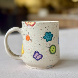 May include: A speckled ceramic mug with a white handle. The mug is decorated with colorful, whimsical shapes, including stars, buttons, and smiley faces. The mug has a slightly textured surface and a rounded base.