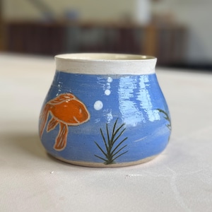 May include: A ceramic, hand-painted fishbowl-shaped vase. The vase features a blue background with an orange goldfish, white bubbles, and green aquatic plants. The rim is white, and the vase has a rounded shape.